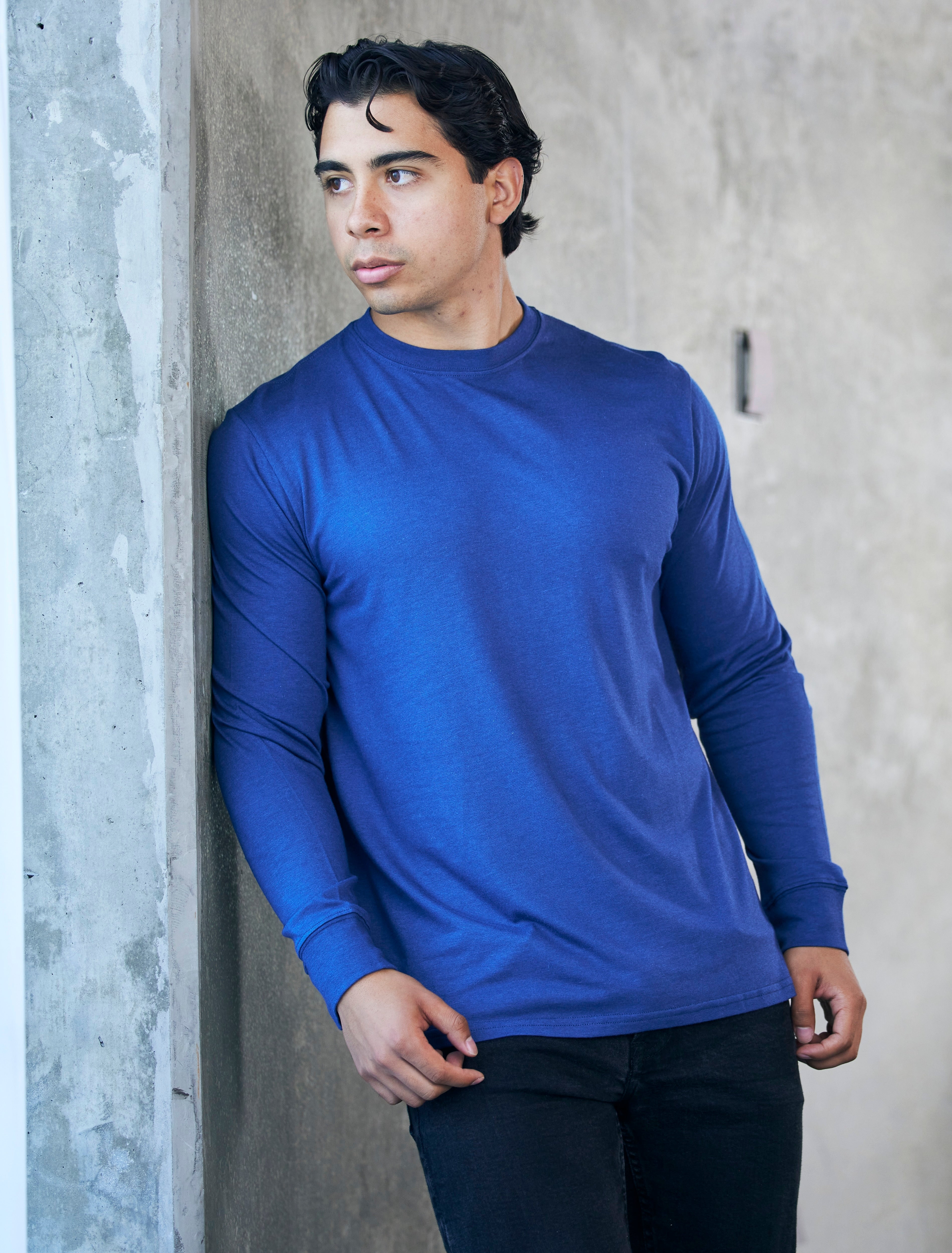 Man wearing a blue long-sleeve shirt leaning against a concrete wall.