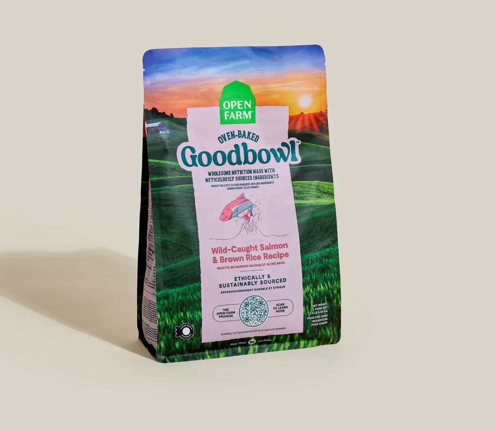 A bag of Open Farm Good Bowl dog food, Wild-Caught Salmon & Brown Rice Recipe, standing upright on a light beige background. The packaging features a scenic landscape of green fields under a sunset sky, with a central white panel displaying the brand name.