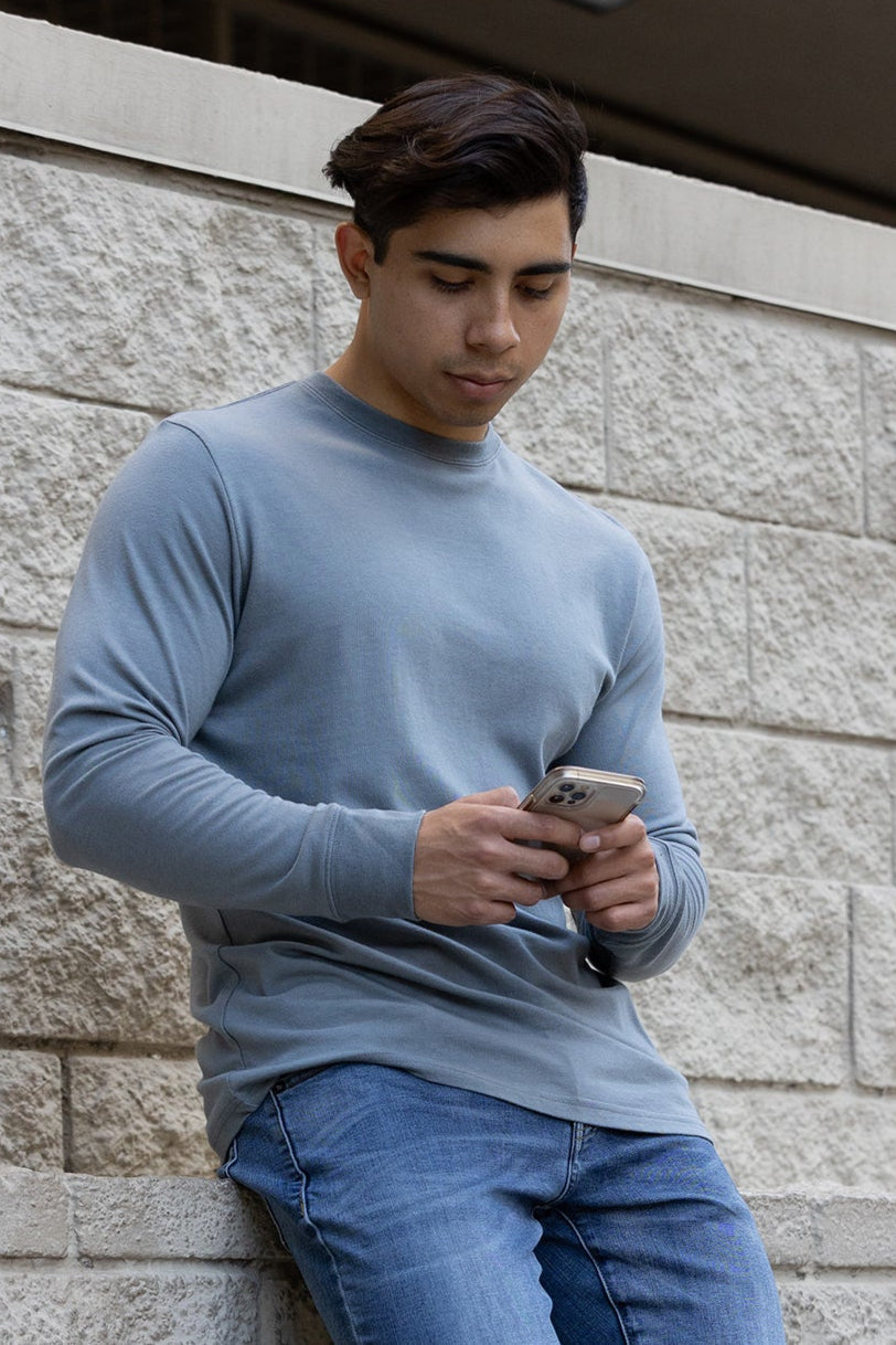 Man in a blue sweater and jeans using a phone against a stone wall.