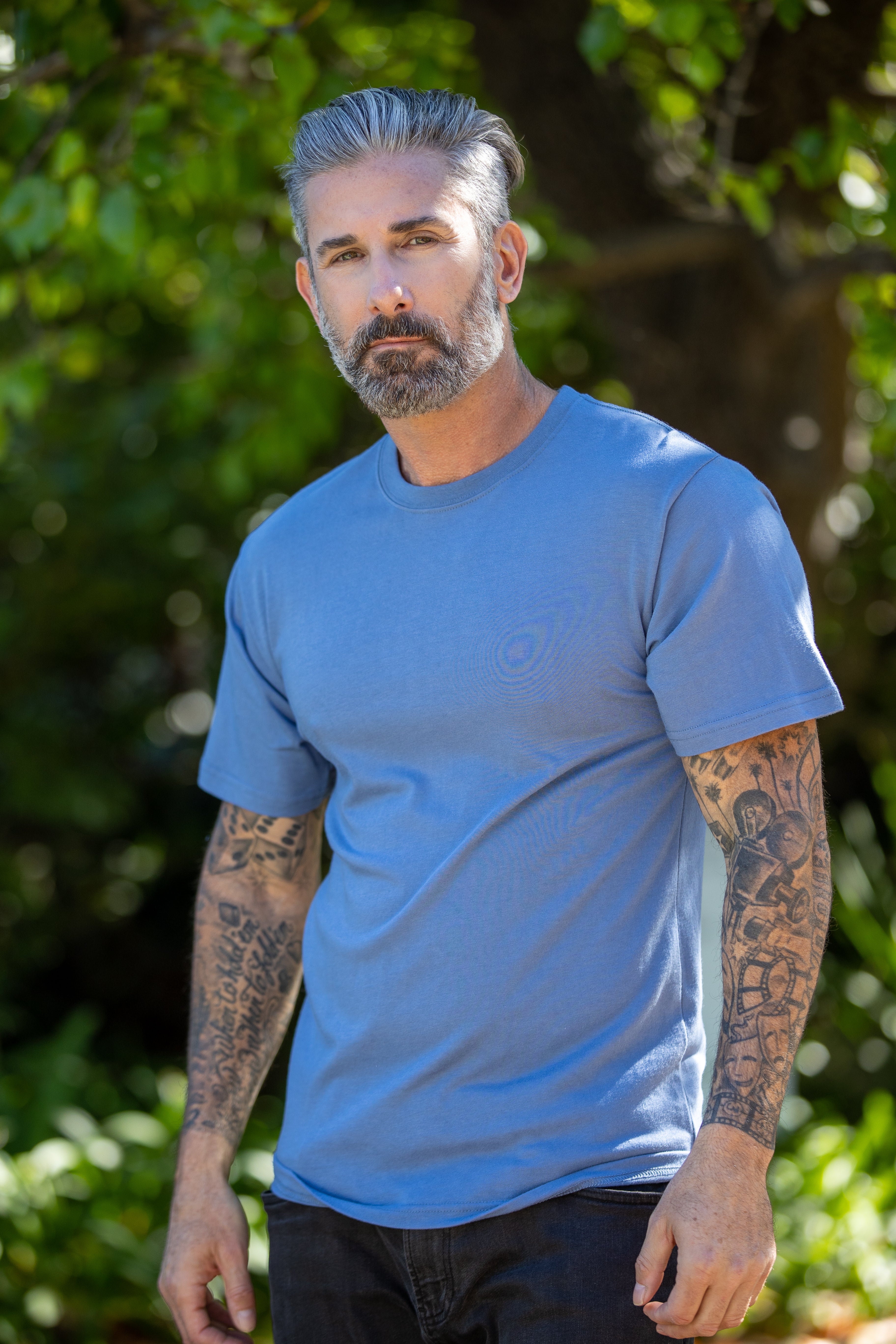 Man wearing a blue t-shirt with tattoos on his arms standing outdoors.